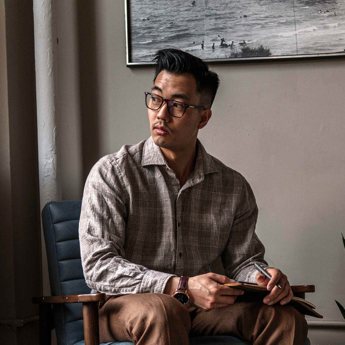 Man sitting in a chair holding a pen and paper, wearing glasses and a checkered shirt.