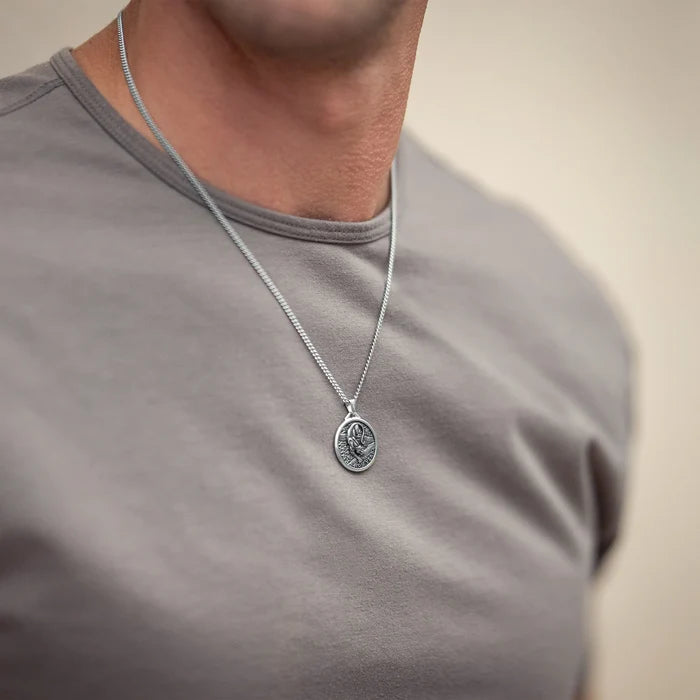 A close-up of a man wearing a silver chain necklace with an oval pendant, paired with a gray t-shirt against a soft beige background.