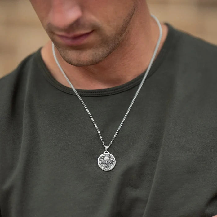A silver chain necklace with a round, coin-style pendant featuring a classical bust, worn by a man in a dark olive green t-shirt.