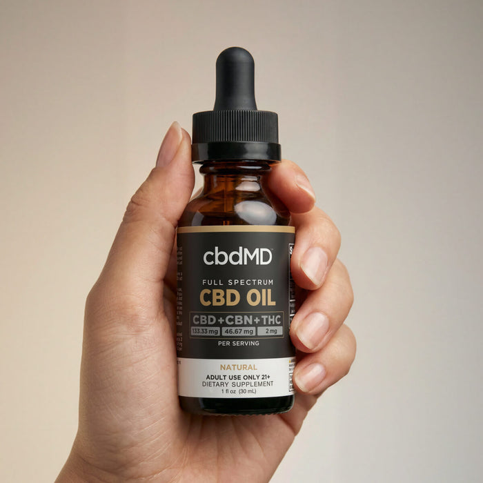 A hand holding a bottle of cbdMD Full Spectrum CBD + CBN Oil tincture with a dropper.