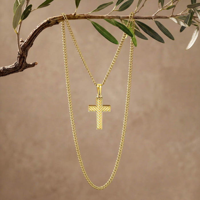 A gold cross pendant necklace hanging from a gold chain, suspended from an olive branch against a warm beige background.