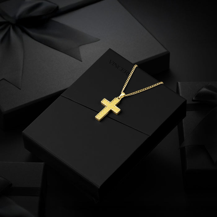 A gold cross pendant necklace on a gold chain, displayed on a black jewelry box with a black ribbon bow, set against a dark background.