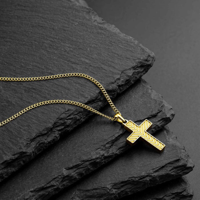 A gold cross pendant necklace with a textured chevron pattern on the cross and a matching chain, displayed on dark textured slate stones.