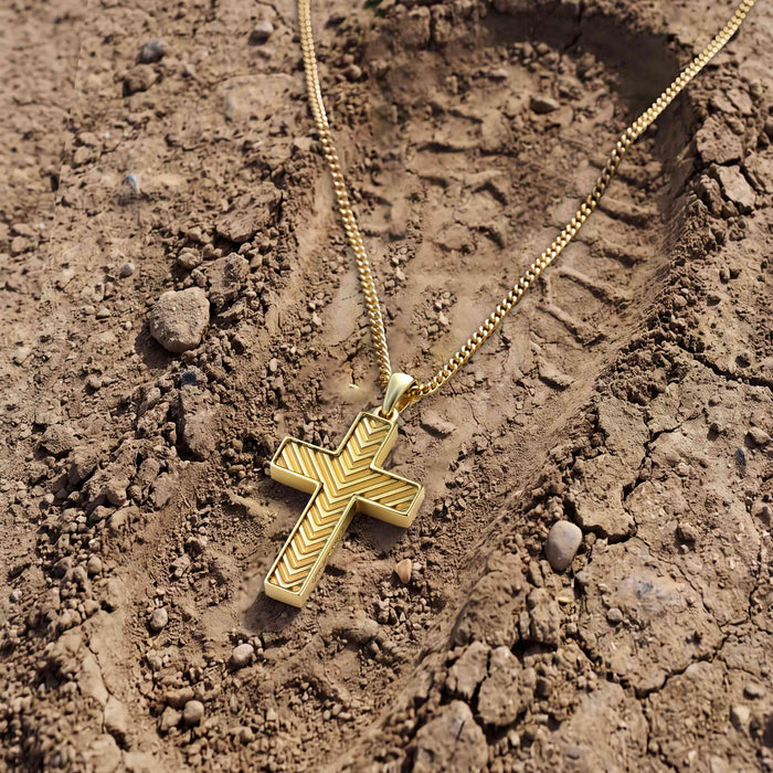 A gold cross pendant necklace with a textured chevron pattern, resting on a dry, cracked brown dirt surface.