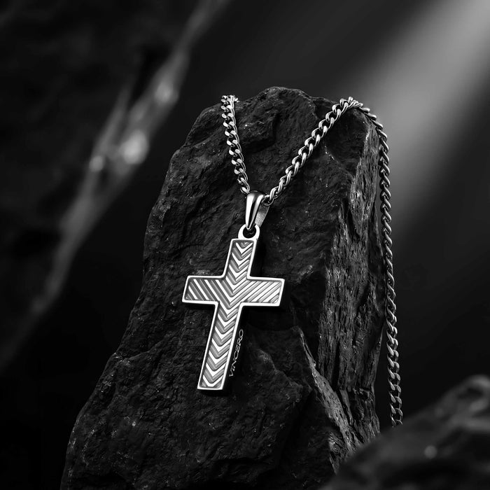 A silver cross pendant necklace with a textured chevron pattern, hanging from a curb chain and resting on a dark rock.