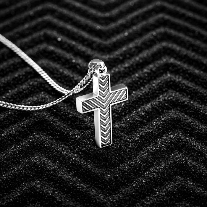 A silver cross pendant necklace with a textured chevron pattern, resting on a black textured background.