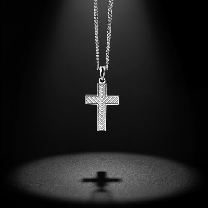 A silver cross pendant necklace with a textured chevron pattern, suspended from a silver chain against a dark background with a spotlight effect and a soft reflection below.