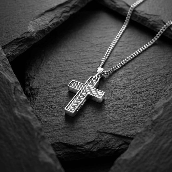 A silver cross pendant necklace with a textured chevron pattern, resting on a dark slate surface.