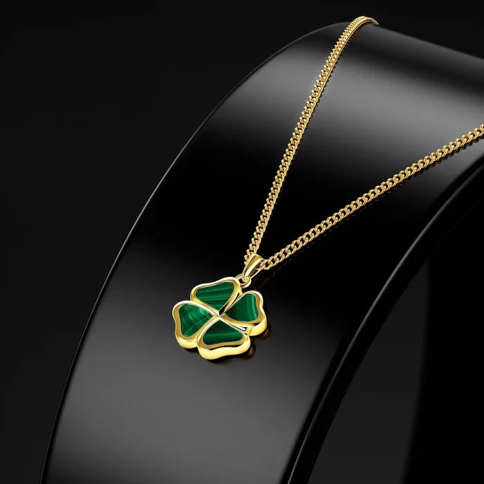 A gold chain necklace with a four-leaf clover pendant featuring green inlay, displayed on a glossy black jewelry bust against a dark background.