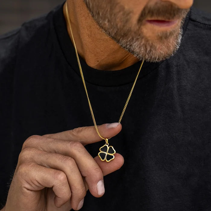 Man wearing a black shirt holding a gold necklace with a clover pendant.