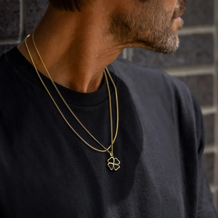 Man wearing a black shirt with a gold necklace featuring a clover pendant against a brick wall.