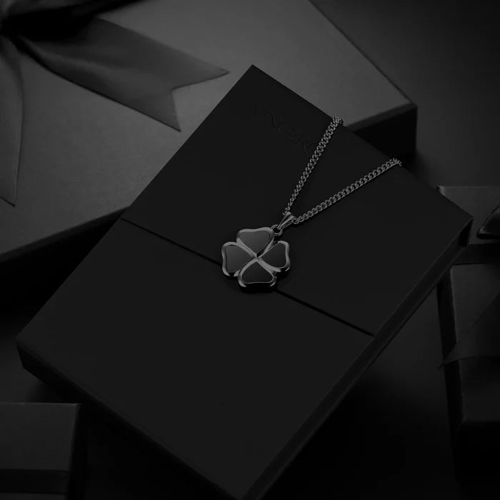 A silver-toned clover pendant necklace displayed on a black presentation box, with dark gift packaging in the background.