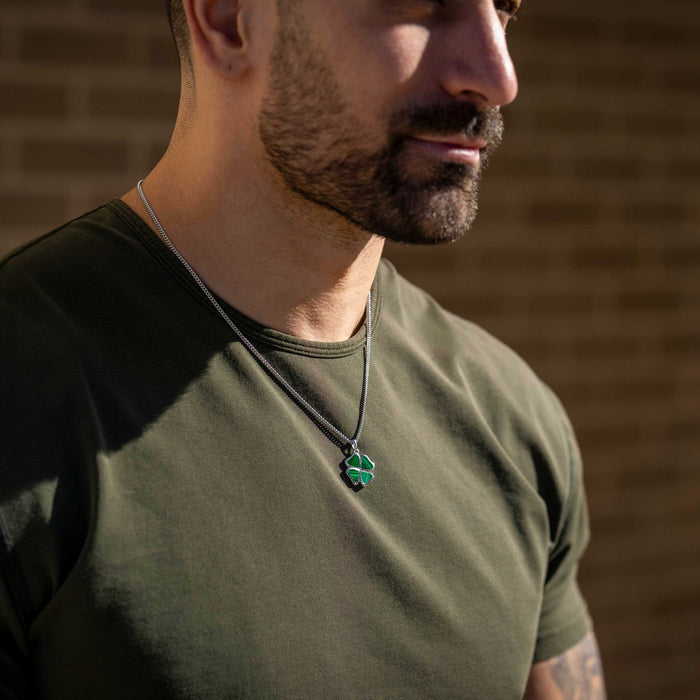 Man wearing a green shirt and necklace with a clover pendant against a brick wall.
