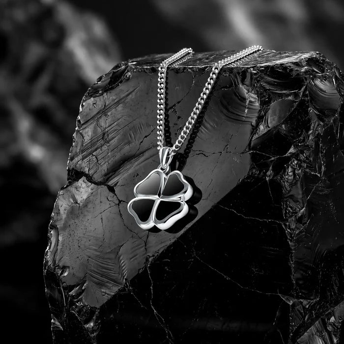 A silver-toned four-leaf clover pendant necklace on a matching chain, displayed on a dark polished rock against a blurred black-and-white background.