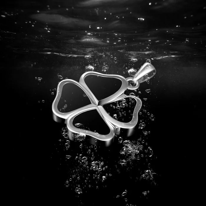 A silver four-leaf clover pendant with an open center, suspended in dark water with visible bubbles and ripples on the surface.
