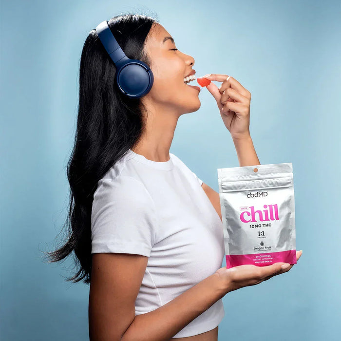 Woman with headphones eating a gummy from a bag labeled chill 10mg THC dragon fruit flavor.