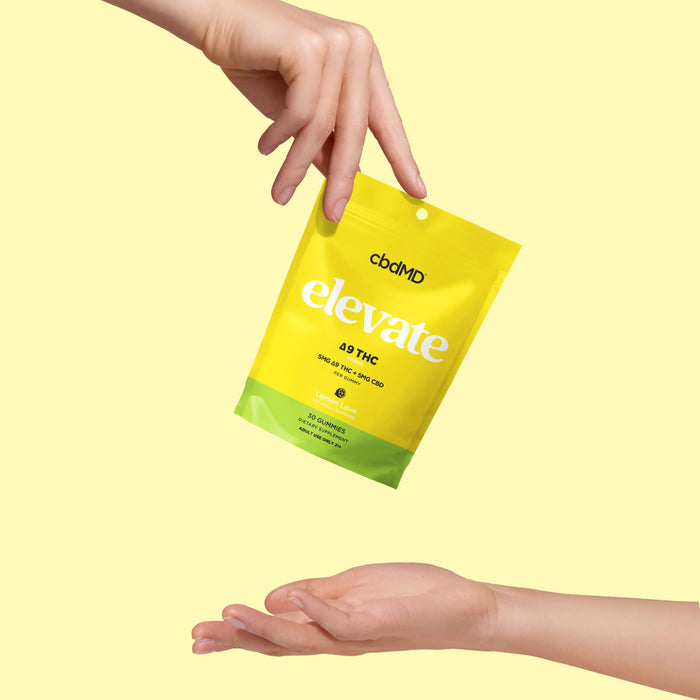 Hand holding cbdMD Elevate Delta 9 THC gummies bag above another open hand on yellow background
