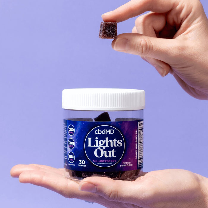 Hand holding cbdMD Lights Out gummies jar with another hand holding a single gummy, purple background