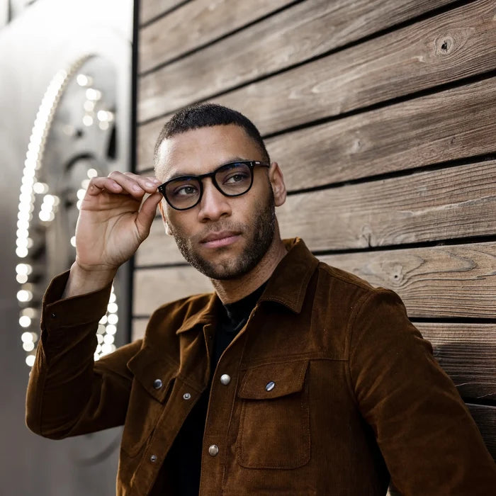 Man wearing glasses and a brown jacket against a wooden wall.