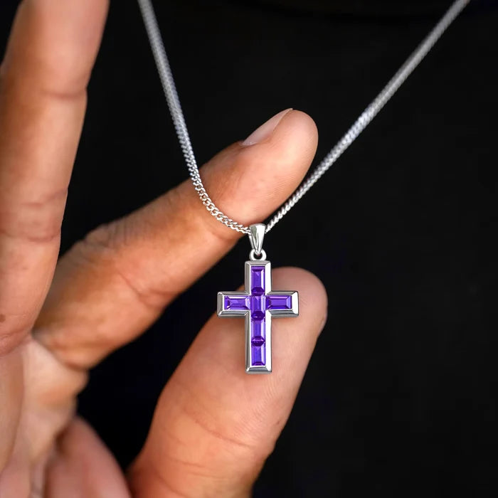 Purple cross pendant on a silver chain held by a hand against a black background