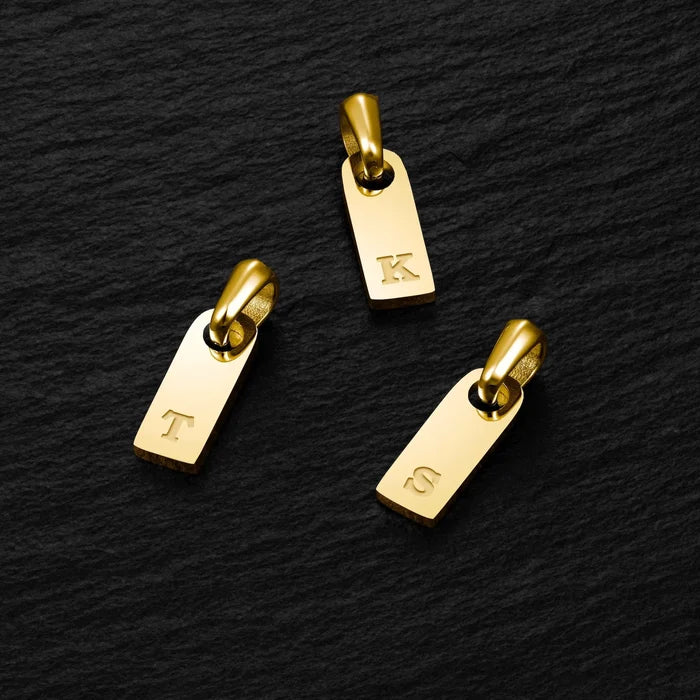 Three gold charms with engraved letters on a black textured background
