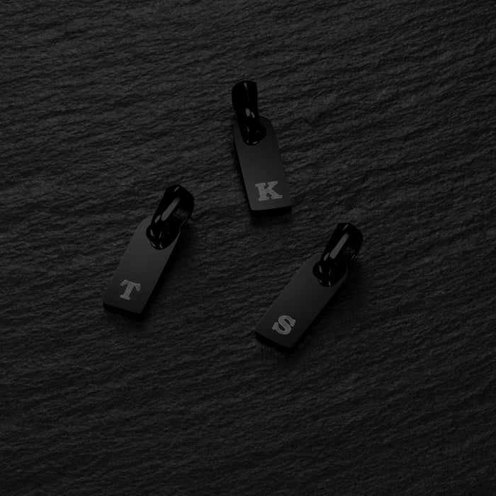 Three black zipper pulls with letters on a dark textured background
