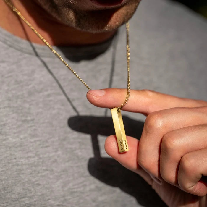 Gold necklace with a bar pendant held by a person wearing a gray shirt.