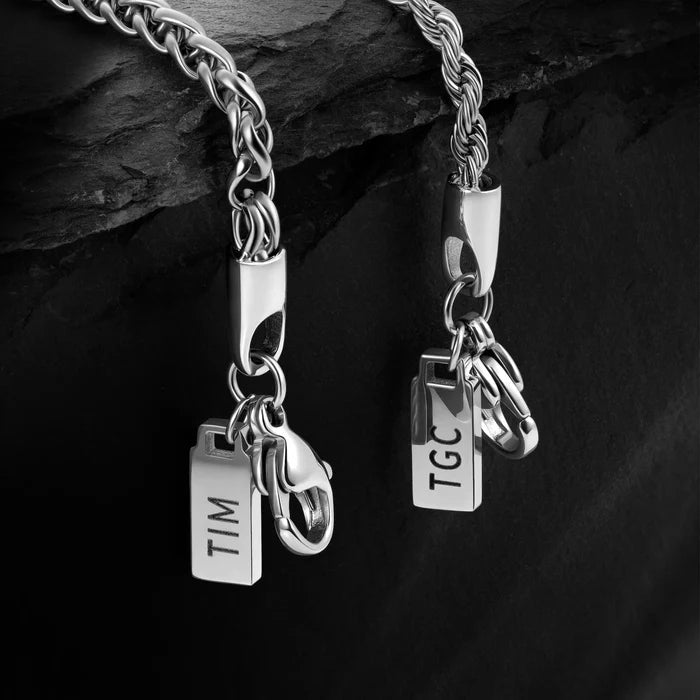 Silver bracelet with two tags, one labeled 'TIM' and the other 'TGC', on a dark stone background.