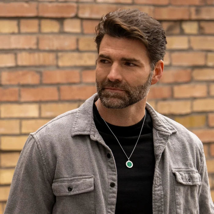 Man wearing a gray jacket and black shirt with a necklace, standing against a brick wall.