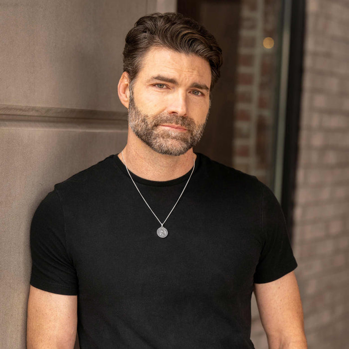 Man wearing a black t-shirt with a necklace against a brick wall.