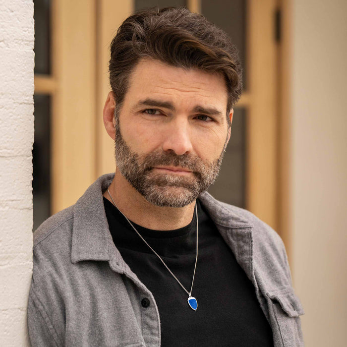 Man wearing a gray jacket over a black shirt with a blue pendant, standing against a neutral background.