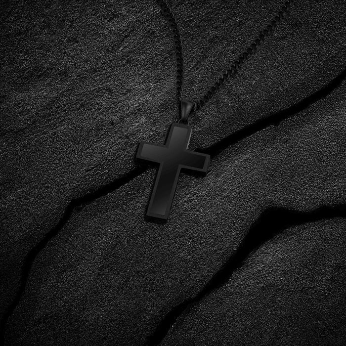 Close-up of a black cross pendant with a black chain, displayed on black sand.