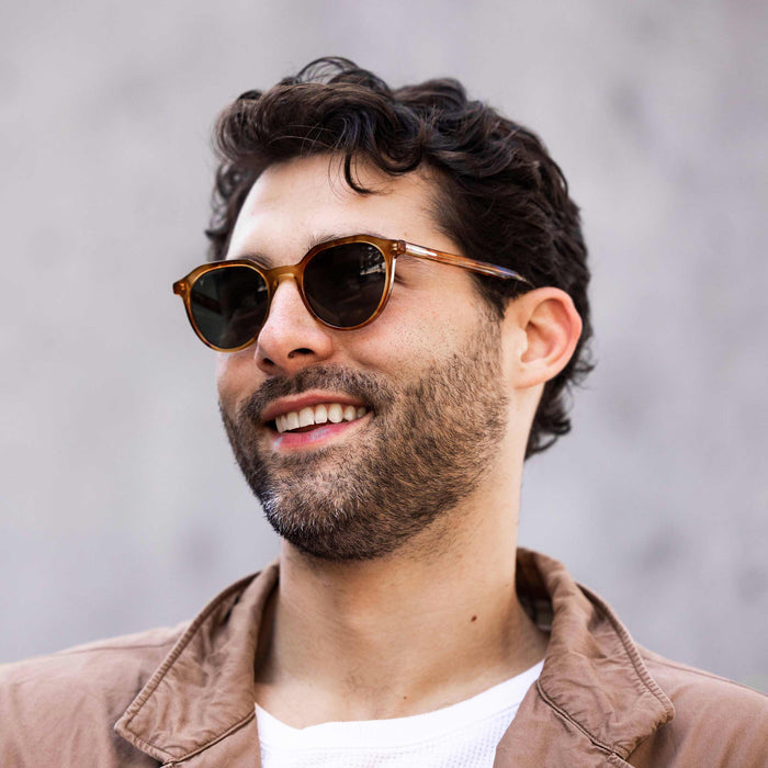 Man wearing sunglasses and a brown jacket against a gray background