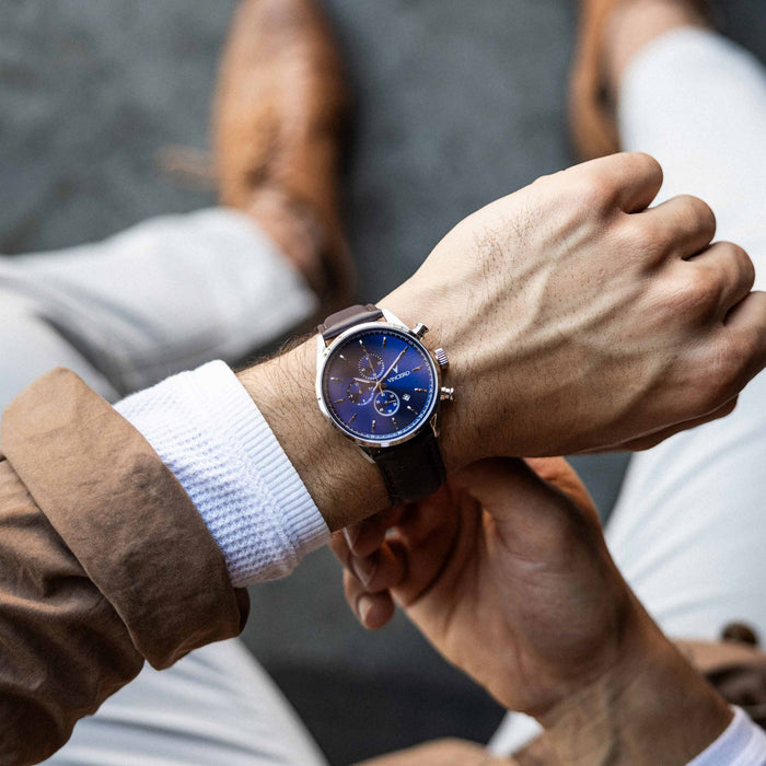 Close-up of two hands with a watch on one wrist, wearing brown jackets and white shirts.