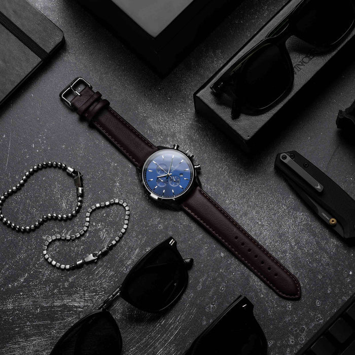 Watch with a brown leather strap on a dark surface with sunglasses and a necklace.