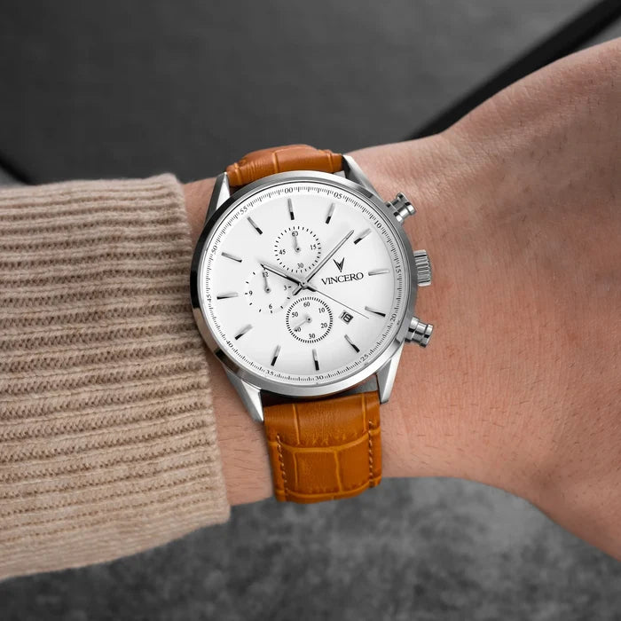 Wrist wearing a watch with a brown leather strap and white dial on a blurred background