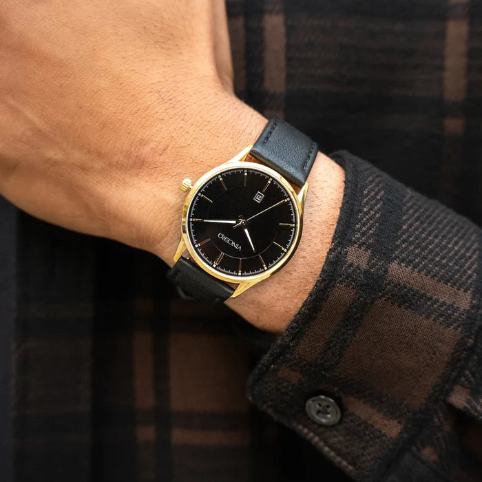 Black watch with gold accents on a wrist, worn with a plaid shirt.