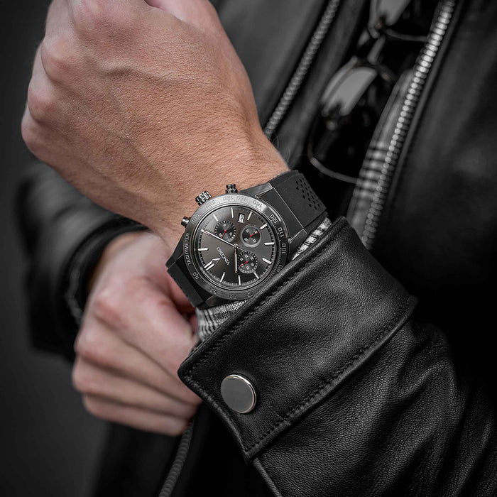 Close-up of a person wearing a black leather watch with a detailed face on a dark background.