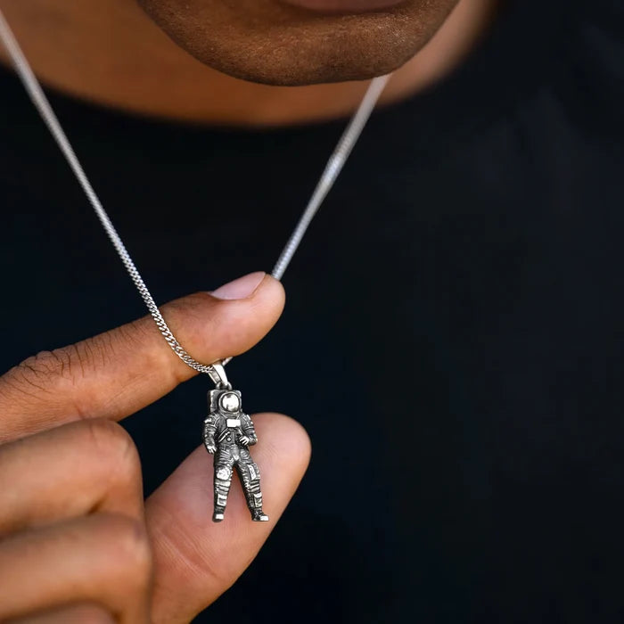 Silver astronaut pendant on a chain held by a hand against a dark background