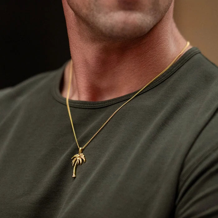 A close-up of a man wearing a gold chain necklace with a small palm tree pendant, paired with an olive green crewneck t-shirt.