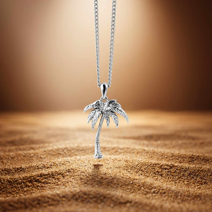 A silver-toned necklace with a detailed palm tree pendant resting on sand against a warm brown gradient background.