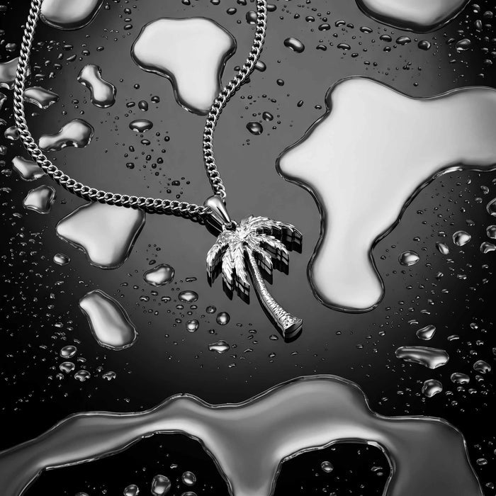 A silver-toned palm tree pendant necklace on a matching chain, photographed on a glossy black surface covered with water droplets and reflective puddle-like shapes.