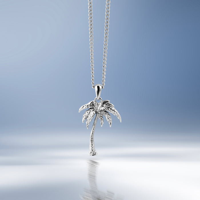 A silver palm tree pendant necklace hanging from a delicate silver chain against a soft blue gradient background with a reflective surface below.